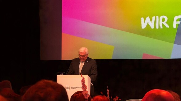 Frank-Walter Steinmeier steht an einem Rednerpult und hält eine Rede. Er schaut nach unten auf das Pult. Hinter ihm wird ein Hintergrund an die Wand gestrahlt, in den Farben: lila, pink, grün, blau.