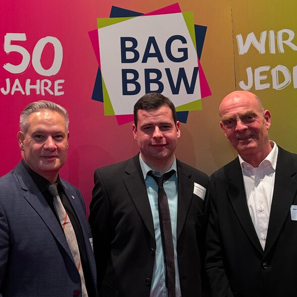 Direktor des BBWs, Alexander Schmidt, ein Teilnehmender sowie ein Mitarbeiter des BBWs stehen nebeneinander und lächeln. Hinter Ihnen ist ein pink-grünes Plakat auf dem steht: 50 Jahre BAG BBW.