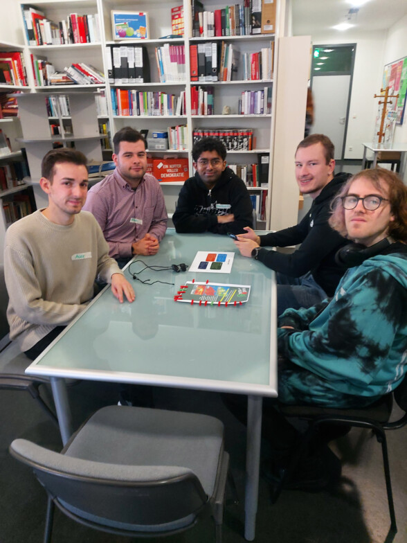 Fünf junge Männer sitzen an einem Tisch und lächeln in die Kamera. Hinter ihnen steht ein Bücherregal.