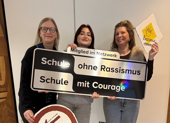 Drei Frauen halten ein schwarz-silbernes Schild hoch. Darauf steht: Mitglied im Netzwerk "Schule ohne Rassismus - Schule mit Courage". Die drei Frauen sind die Bezirksrätin und Patin des Projekts der Berufsschule, eine Schülerin und eine Lehrerin.