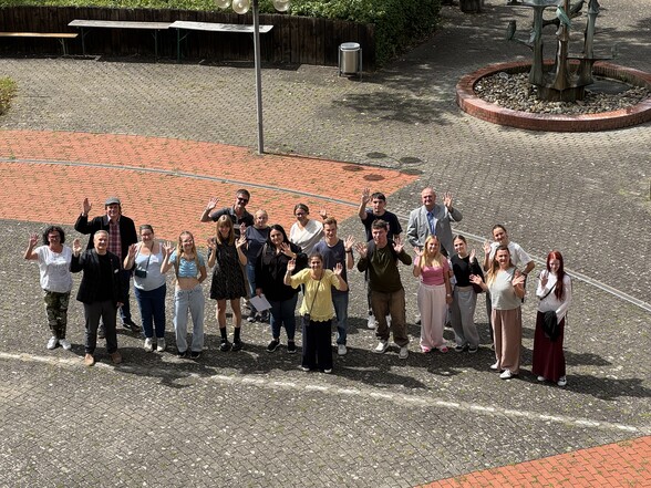 Rund 20 Menschen stehen zusammen auf dem Pausenhof des BBW und schauen nach oben in die Kamera und winken. 