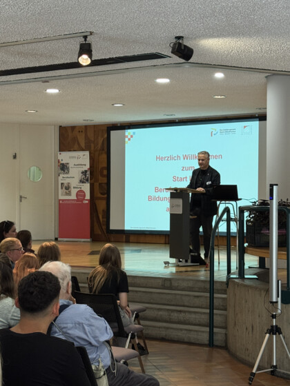 Der Direktor des Berufsbildungswerks, Herr Schmidt, steht auf der Bühne in der Aula hinter einem Rednerpult. Vor der Bühne sitzen mehrere junge Erwachsene.