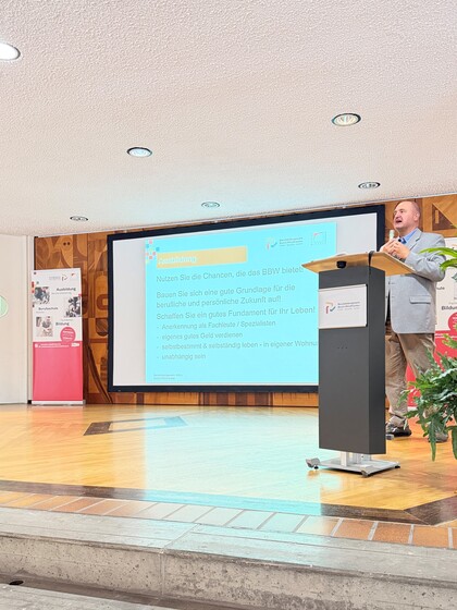 Der Ausbildungsleiter Herr Klaus steht auf der Bühne hinter einem Rednerpult. Er trägt einen Anzug und hält ein Mikrofon in der Hand. Hinter ihm wird eine Power-Point-Präsentation an die Wand projiziert mit der Überschrift "Ausbildung".