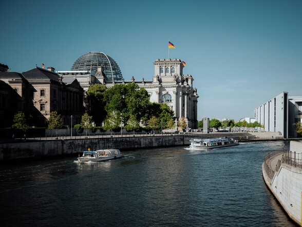 Im Vordergrund ist die Spree mit zwei Fahrgastschiffen. Im Hintergrund ist teilweise der deutsche Bundestag zu sehen, leicht verdeckt von Bäumen.