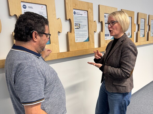 Eine Frau und ein Mann stehen vor Informationsstafeln. Sie schauen sich gegenseitig an und die Frau gebärdet.
