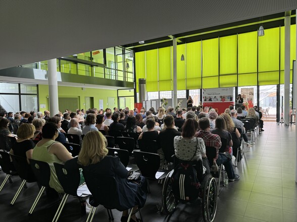 Rund 80 Menschen sitzen in Stuhlreihen vor einer Bühne. Vorne rechts steht auf der Bühne eine Frau hinter einem Rednerpult. Links von ihr steht eine Gebärdensprachdolmetscherin.