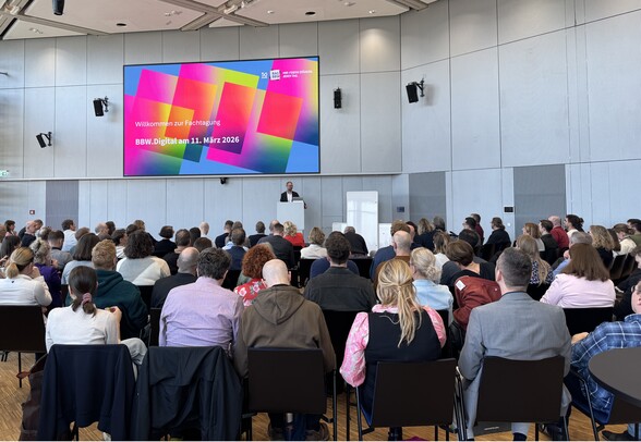Auf einer Bühne steht ein Mann hinter einem Rednerpult und spricht. Hinter ihm hängt ein großer Bildschirm, darauf steht: Willkommen zur Fachtagung BBW.Digital am 11.März 2026. Man sieht etwa 60 Personen von hinten, die im Publikum sitzen.